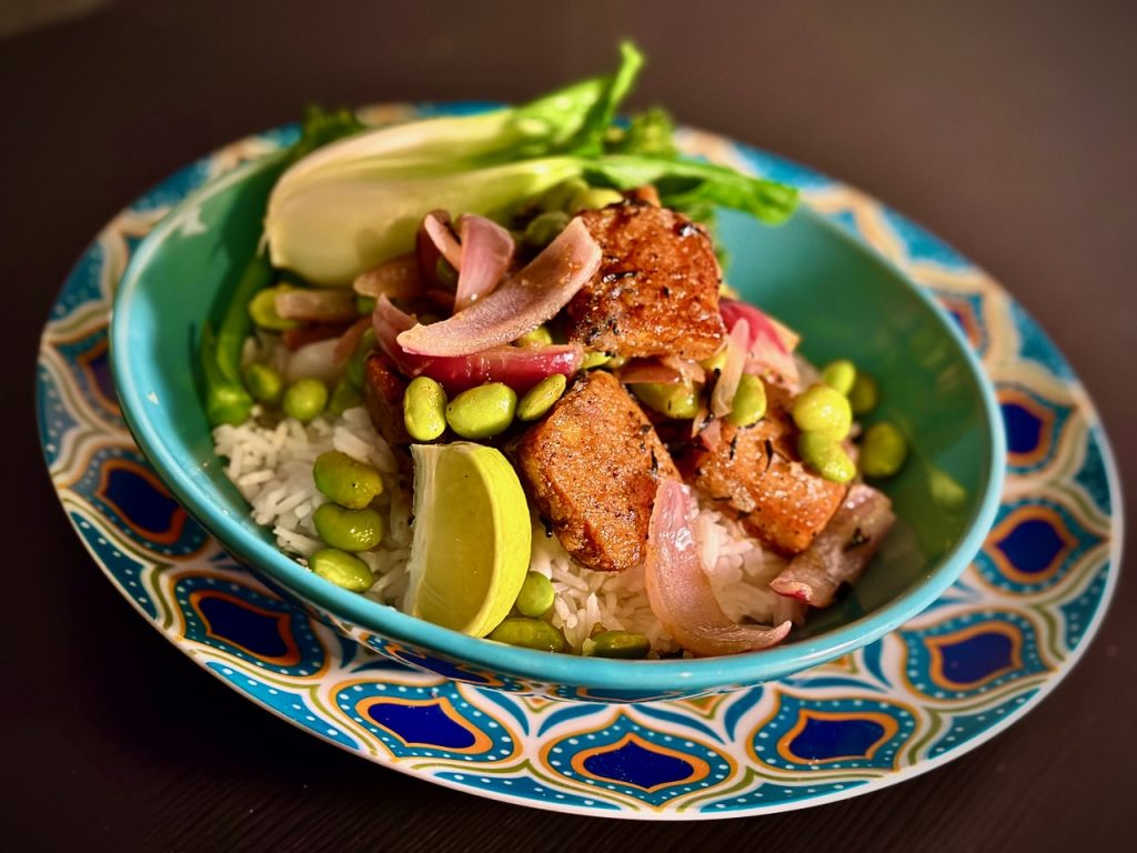 Turquoise bowl with pan-friend tofu on top of white basmati rice, with green edamame beans, red onion petals and a lime wedge.