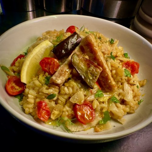 White bowl with bright and colour risotto with tomatoes and herbs, a lemon wedge, some chunks of purple aubergine and thick chunky tempeh chips