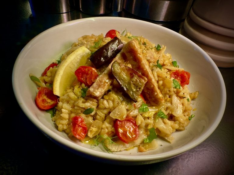 White bowl with bright and colour risotto with tomatoes and herbs, a lemon wedge, some chunks of purple aubergine and thick chunky tempeh chips