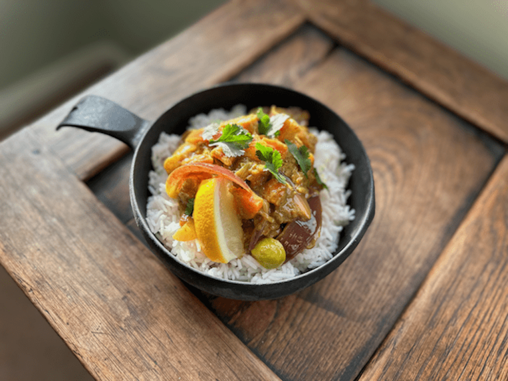 Black bowl of curry on top of white rice with a lemon wedge, topped with fresh coriander