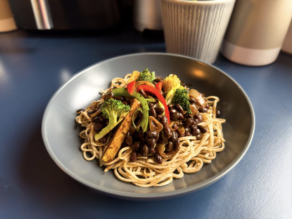 Dark grey bowl filled with wholewheat noodles topped with vegan black bean sauce, red pepper, green pepper, broccoli, and tofu.