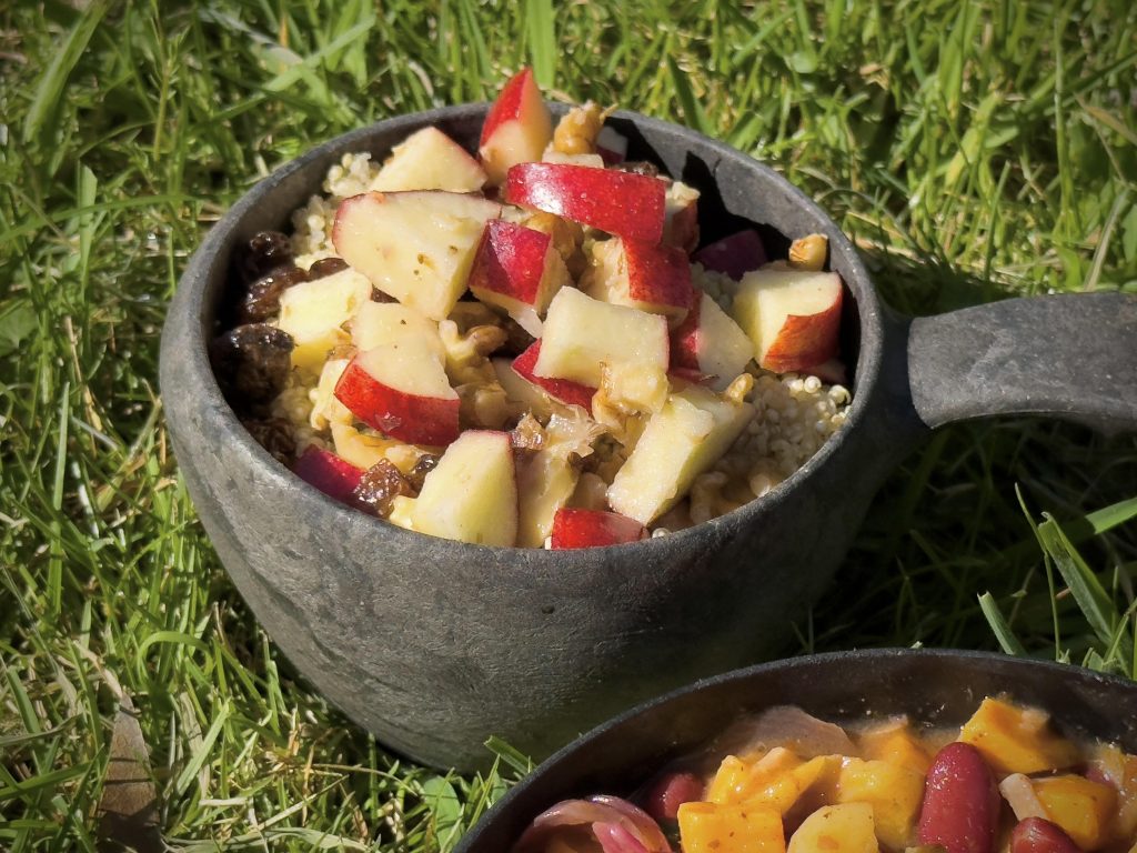 Black bowl outside on green grass filled with a salad made of red skinned apple, walnuts and sultanas