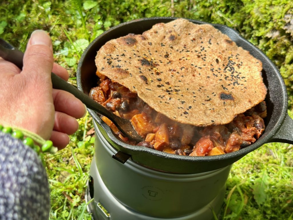 trangia stove with a black skillet pan of vegan sweet potato and black bean chilli, topped with homemade paratha. shows a person's hand stirring.