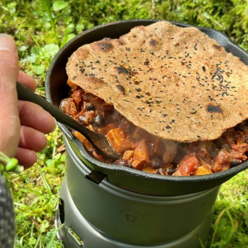 trangia stove with a black skillet pan of vegan sweet potato and black bean chilli, topped with homemade paratha. shows a person's hand stirring.