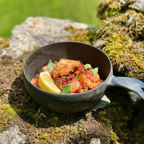 A serving of sticky barbecue beans and golden cornbread dumplings, with herbs and lemon zest scattered on top. a lovely vegan evening meal. It is on a stone wall with mossy around. A gorgeous summer bowl.