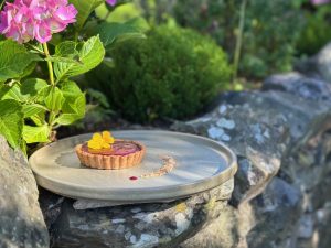 Individual tart filled with deep purple vegan damson curd pictured on a stone wall with flowers around in late evening sun