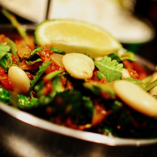 A close up of a butterbean, spinach and tomato curry in a stainless steel indian bowl with a slice of lime on top