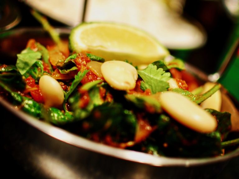 A close up of a butterbean, spinach and tomato curry in a stainless steel indian bowl with a slice of lime on top