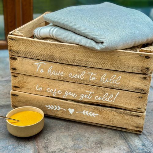 Bowl of red pepper bisque soup in front of lovely decorated wooden crate filled with blankets. A warm cosy image.