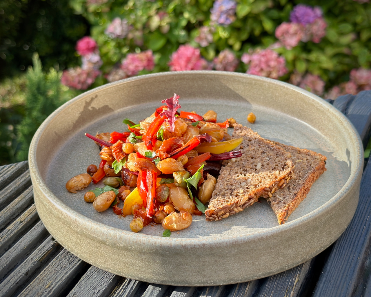 A rustic stoneware bowl containing a delicious fresh vegan roasted chickpeas and red pepper salad. There are slices of wholesome seedy bread. A