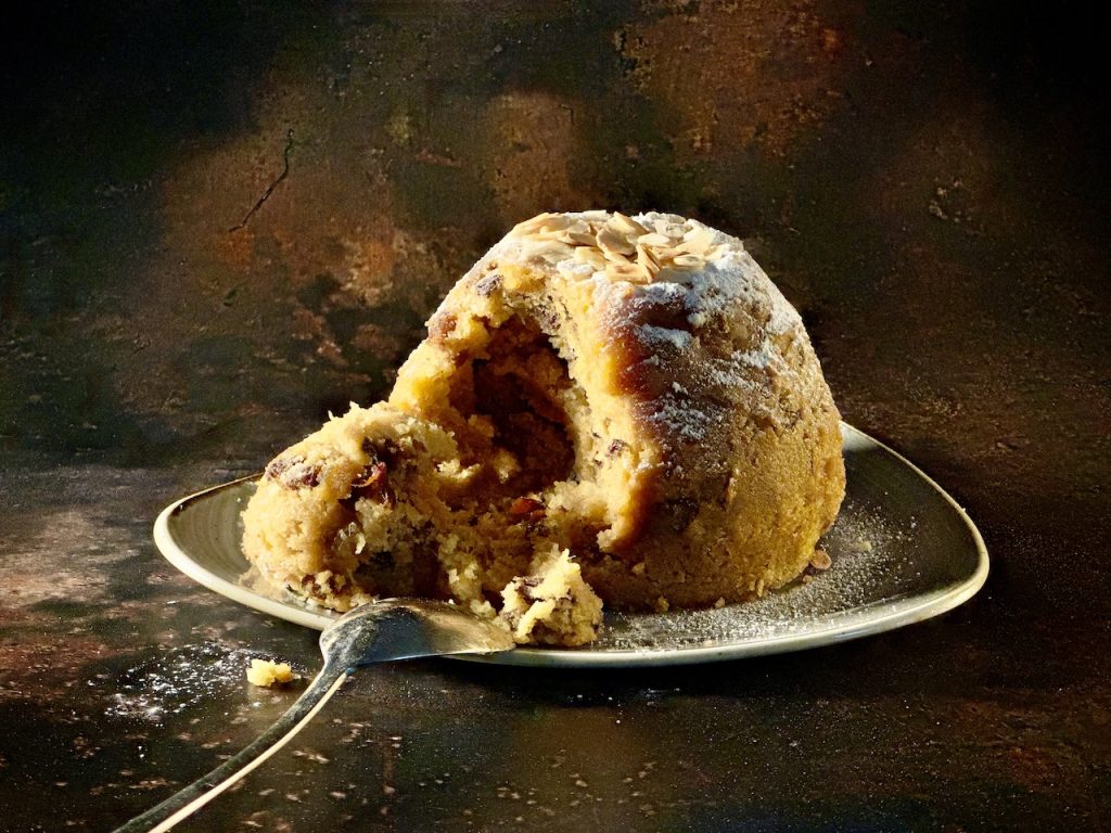 a whole christmas pudding on a plate; the pudding is light in colour, like stollen and is studded with fruit and marzipan. there is lots of icing sugar and toasted almonds